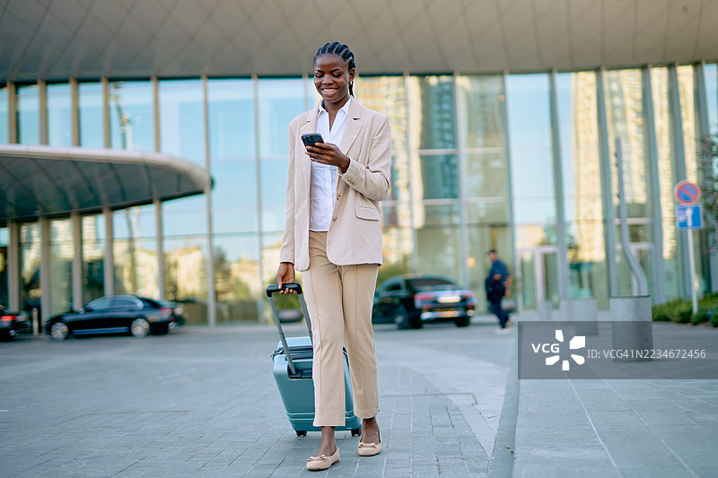 微笑的女商人提着行李箱，边走边使用智能手机，靠近现代建筑。图片素材