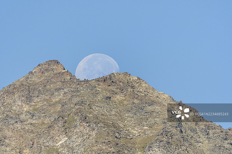 雄伟的山峰在生动的蓝天映衬下高耸,巨大的月亮正从山后升起。这个场景捕捉了傍晚时分自然的宁静之美。瑞士瓦莱州的格拉肯,维耶格。图片素材
