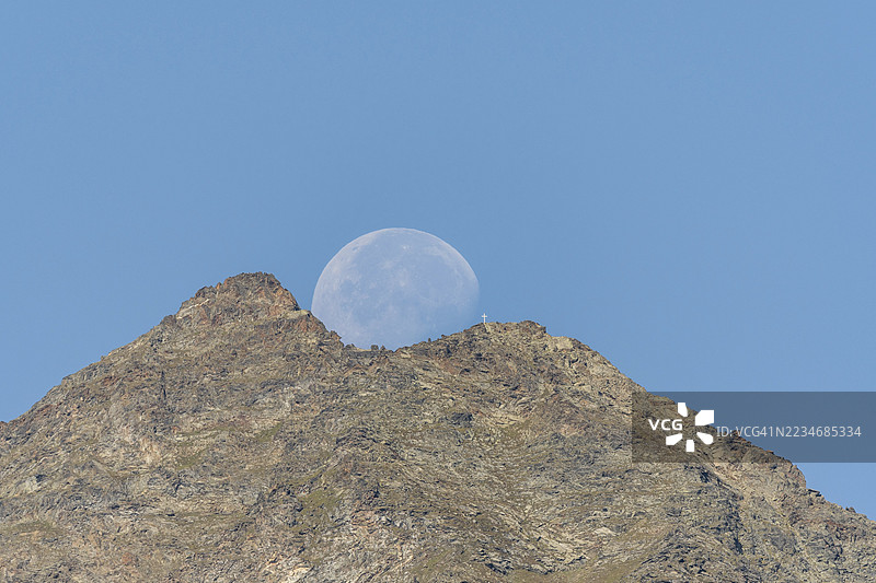 雄伟的山峰在生动的蓝天映衬下高耸,巨大的月亮正从山后升起。这个场景捕捉了傍晚柔和光线下自然的宁静之美。瑞士瓦莱州维耶格的格拉岑。图片素材