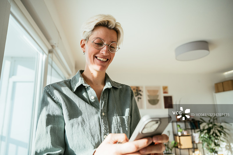微笑的短发金发人士戴着眼镜，在明亮现代的客厅里使用智能手机，周围满是植物。图片素材