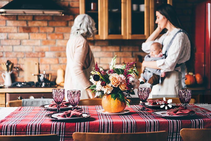 一张布置精美的餐桌，铺着格子桌布，中央摆放着南瓜装饰，背景中模糊地可以看到一位母亲、祖母和婴儿。这张图片强调了多代同堂的家庭聚会、秋季盛宴和节日室内装饰。图片素材