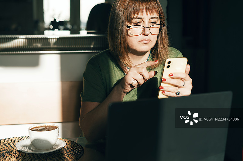 女性在咖啡馆桌子上使用智能手机，同时在笔记本电脑上工作图片素材