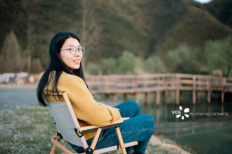 一位年轻的亚洲女性坐在露营椅上，抬头仰望天空，面带微笑。图片素材