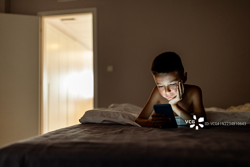 年轻男孩在床上躺着专注于使用智能手机，夜晚时分图片素材