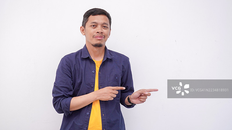 微笑的年轻亚洲男子在白色背景上指向右侧。英俊的男子穿着深蓝色衬衫。图片素材