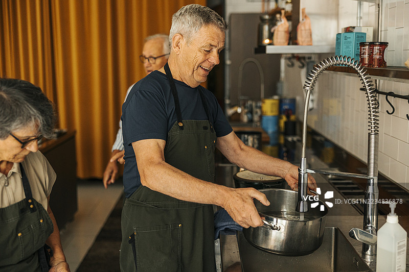 微笑的老年男子在烹饪课上用水龙头的水给炖锅加水，站在厨房水槽旁。图片素材