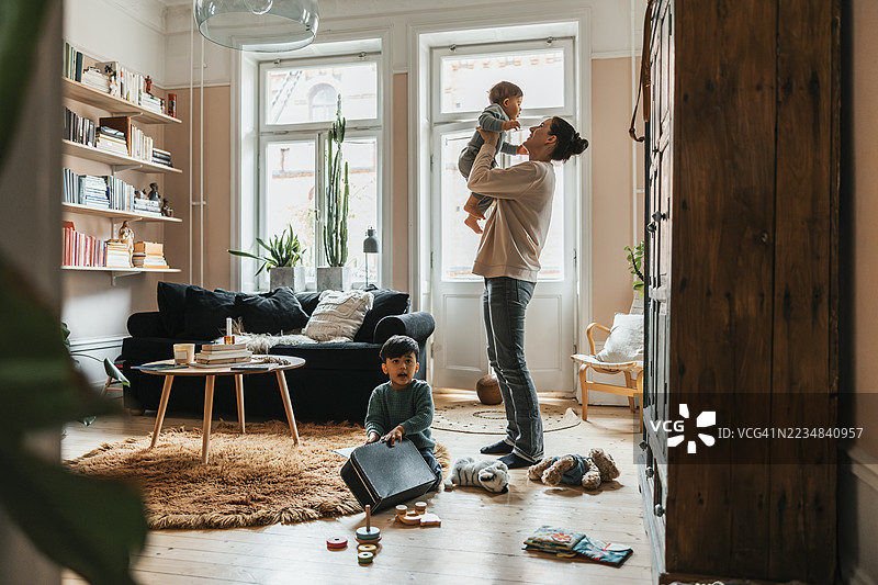 男孩在家里的客厅里玩玩具，母亲正在抱起儿子。图片素材