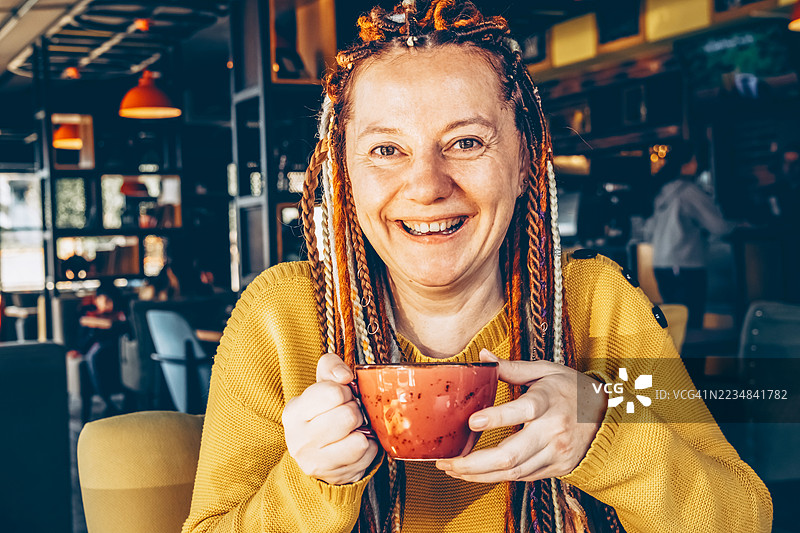 微笑的女性扎着辫子，穿着黄色毛衣，手里拿着红色咖啡杯。温馨咖啡馆里的明亮氛围。图片素材