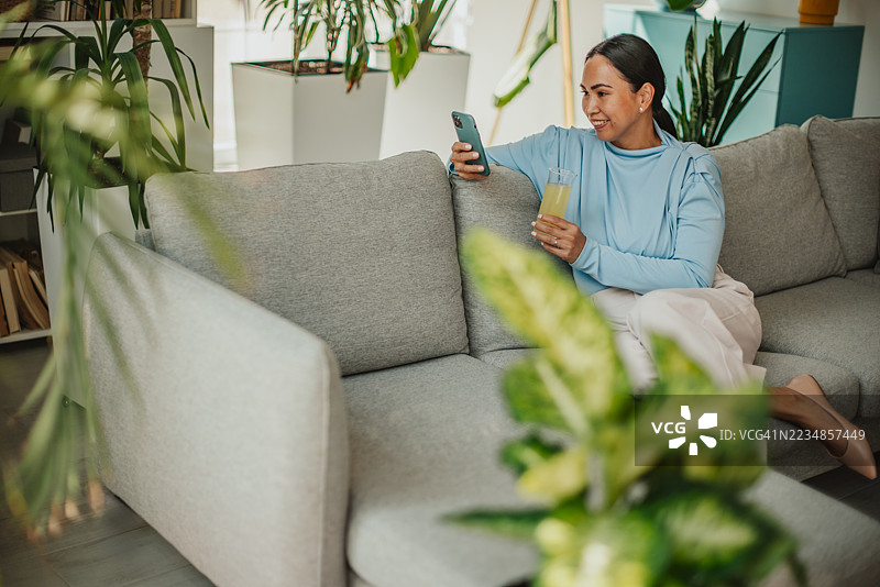 快乐的亚洲女性在家中享受放松时光，手里拿着饮料和智能手机，周围环绕着室内植物。图片素材