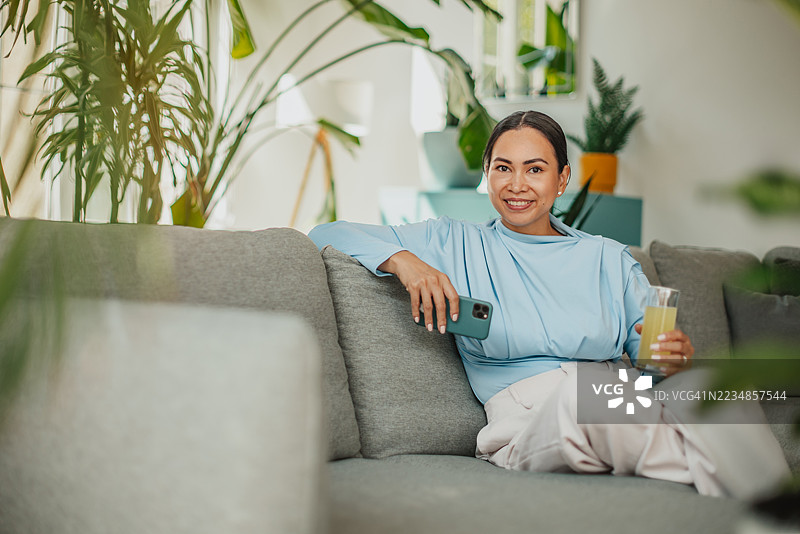 愉快的亚洲女性在现代公寓的舒适沙发上放松，手里拿着饮料和手机，享受宁静的周末时光。图片素材