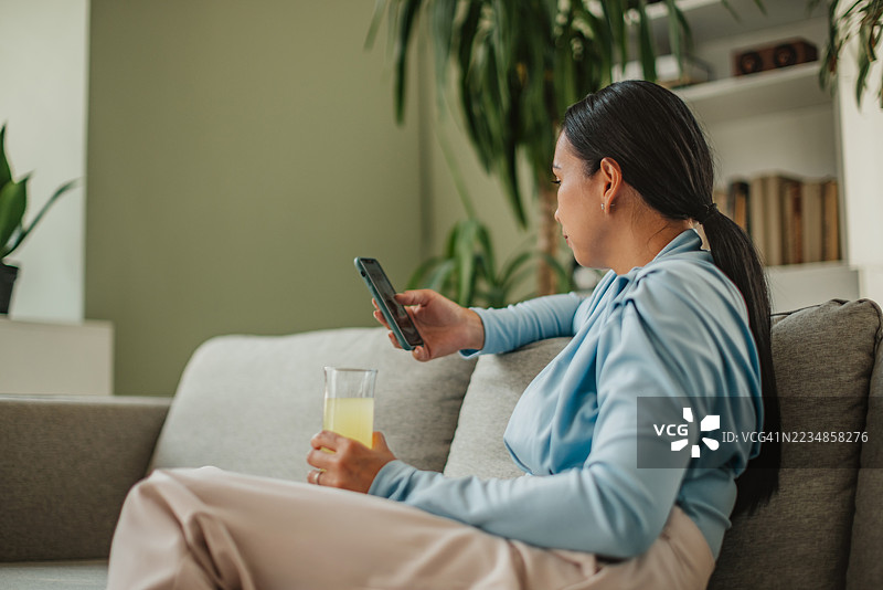 亚洲女性在家中享受宁静时光，手中拿着饮品和手机，度过放松的周末时光。图片素材
