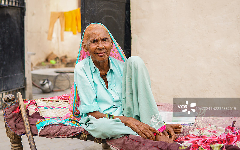 传统的印度农村老年女性坐在查尔派上，面带微笑看向镜头。图片素材