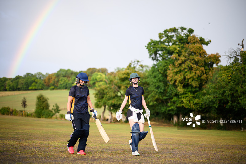 全身镜头拍摄的女板球运动员走出板球场，身后是多云天空中的彩虹。图片素材