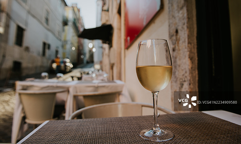 在古镇狭窄街道上的温馨户外咖啡馆桌子上放着一杯白葡萄酒，夕阳西下。图片素材