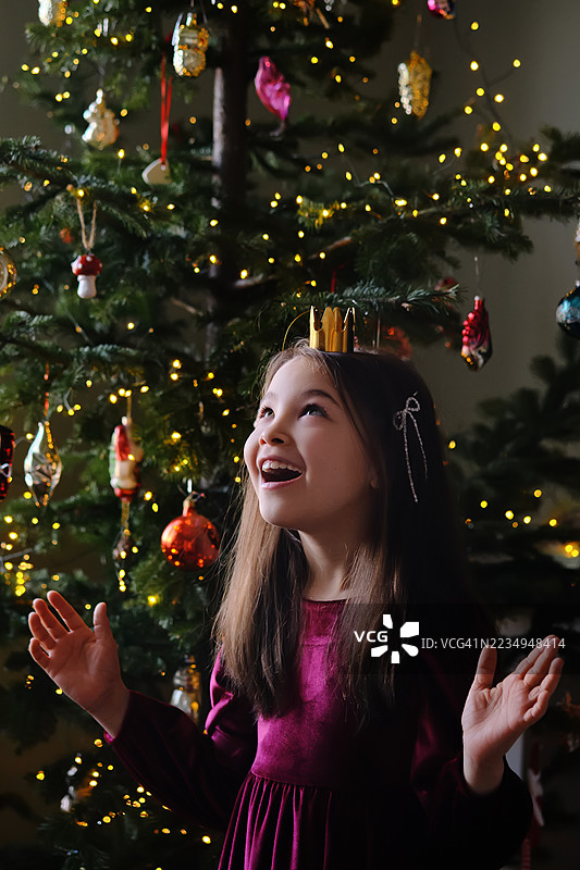 一个穿着酒红色裙子的可爱女孩在装饰好的圣诞树背景下试戴圣诞树皇冠玩具。搞笑的新年照片。图片素材
