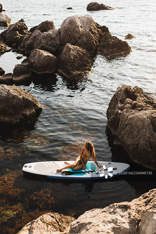 日落时分，女人在岩石海岸水域的立式划水板上放松。图片素材