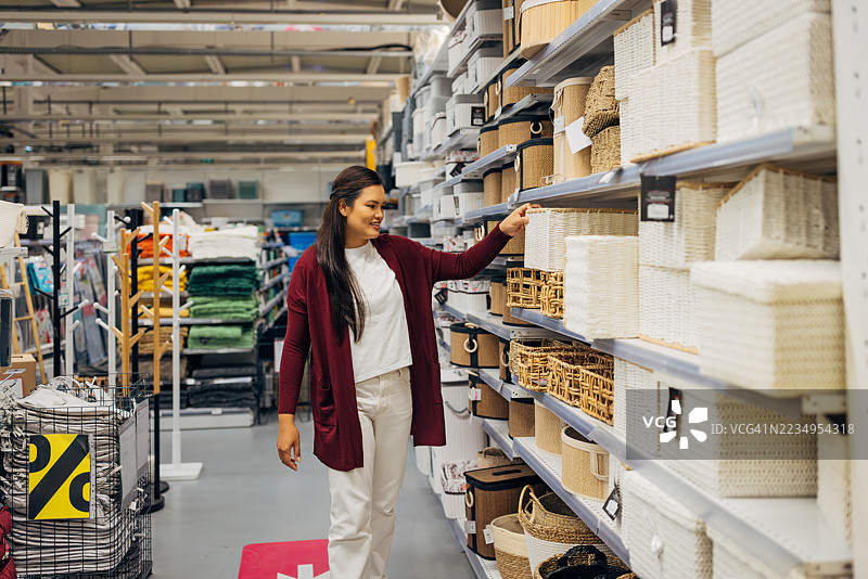 亚洲女性顾客在室内装饰商店内走过整齐排列的储物篮和盒子架子，寻找时尚的家居储物方案图片素材