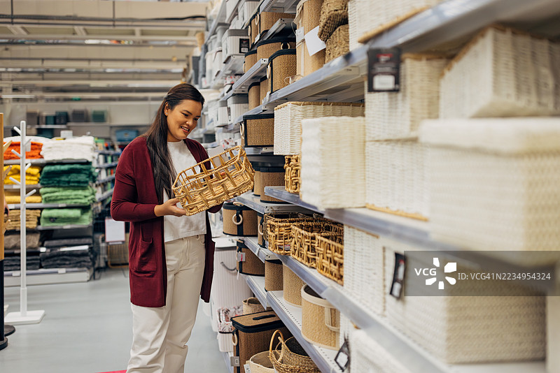 一位亚洲女性在家居商店的货架间走动，查看各种储物篮和箱子。图片素材