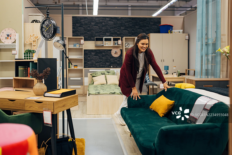 无忧无虑的亚洲女性在卧室主题的家具展厅中检查沙发。沙发上搭配了鲜艳的黄色装饰枕头，为中性色调的室内增添了一抹亮色。图片素材
