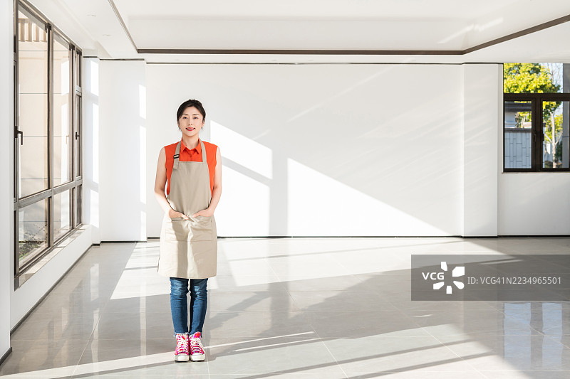 亚洲年轻女性穿着围裙，站在她的新家客厅里，家具将在未来搬入房子。图片素材