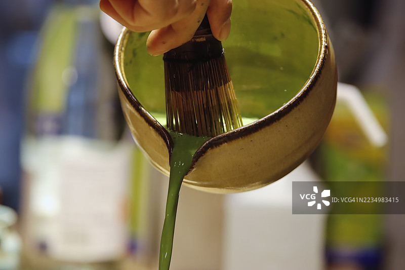 手部特写，正在将浓厚鲜艳的绿色抹茶茶倒出，来自传统陶瓷碗，强调健康日本饮品的丰富质感和制作过程。图片素材