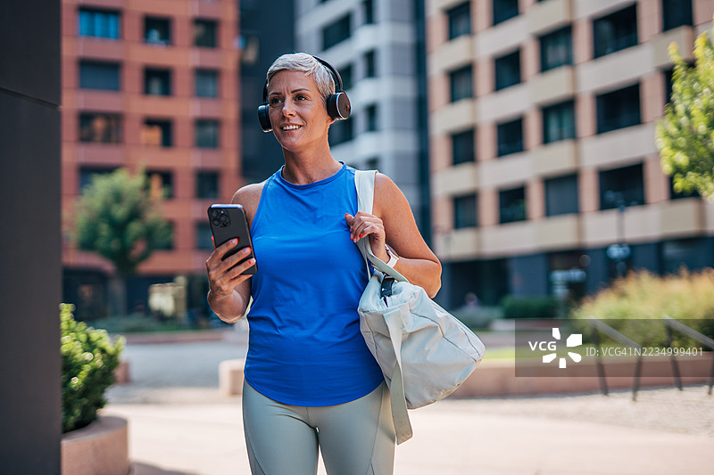 微笑的年轻女运动员在城市中走路，戴着耳机，手持智能手机图片素材