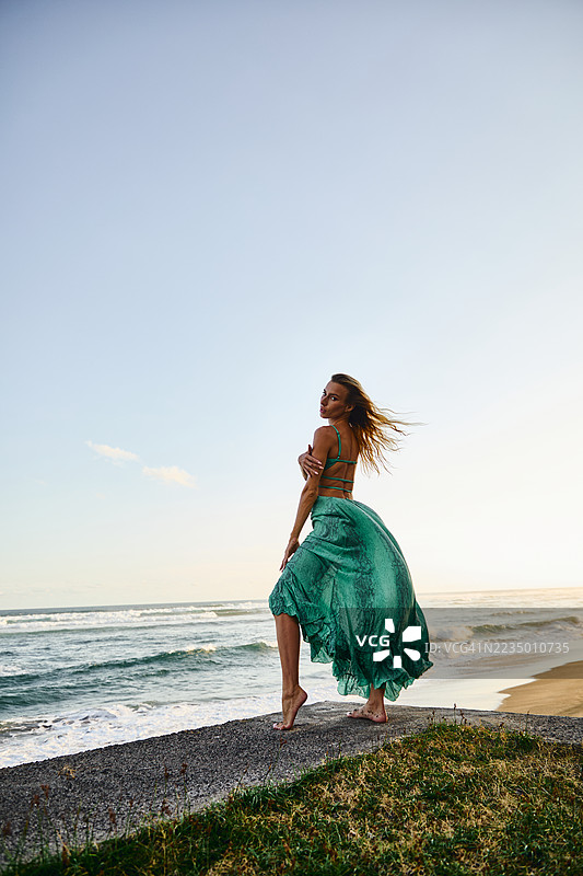 穿着飘逸绿色裙子的女人站在海边悬崖上，夕阳西下。图片素材