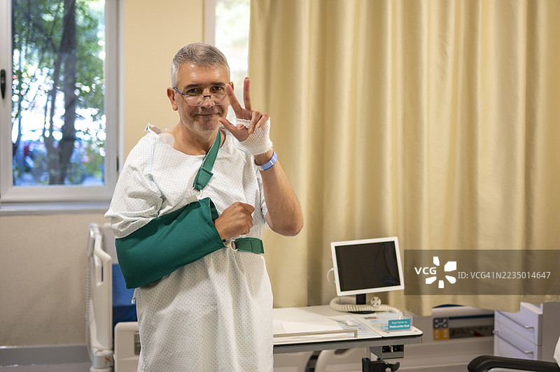 穿着医院病服的成年男性患者，戴着绿色手臂吊带，微笑并做出和平手势。图片素材