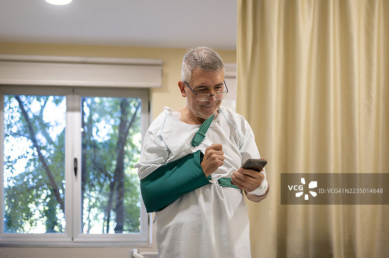 老年患者站在窗边，手臂挂着绿色吊带，正在阅读智能手机。图片素材