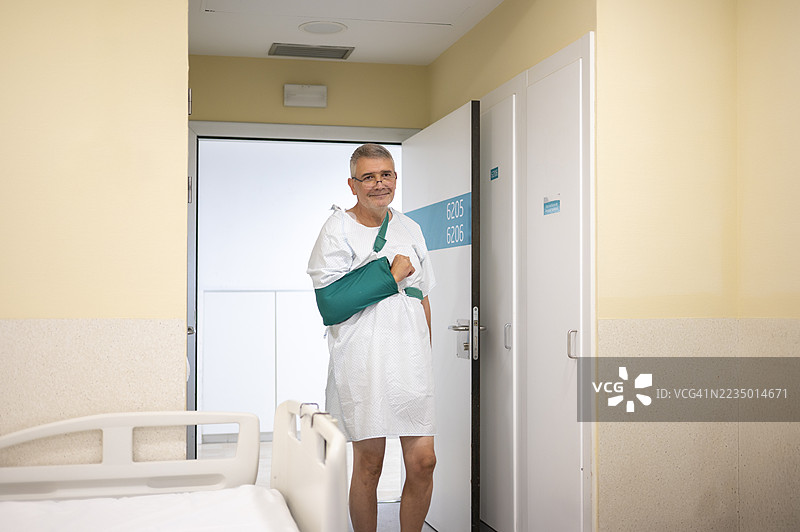 患者微笑着站在医院病房的门口，穿着病号服和绿色吊带。图片素材