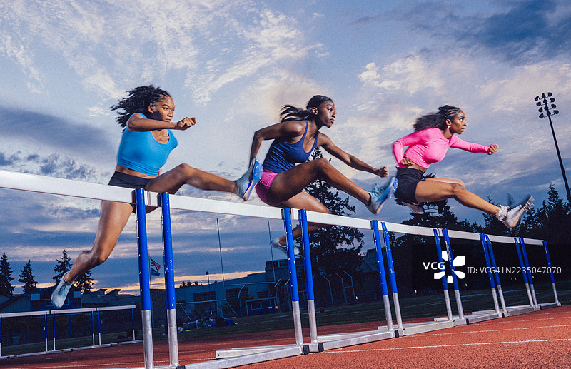 三名运动员穿着鲜艳的运动服在跑道上跳过障碍，背景是美丽的天空。图片素材
