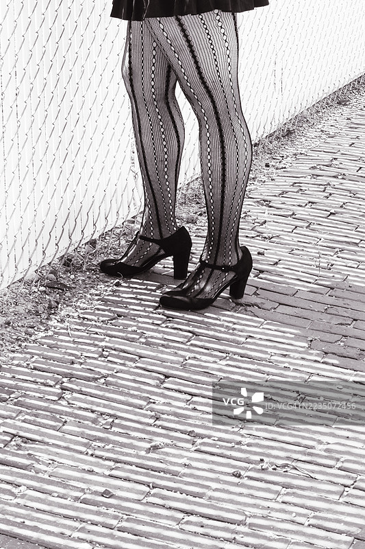 穿着图案丝袜和鞋子的女性下半身站在有纹理的人行道上，旁边有一个围栏。图片素材