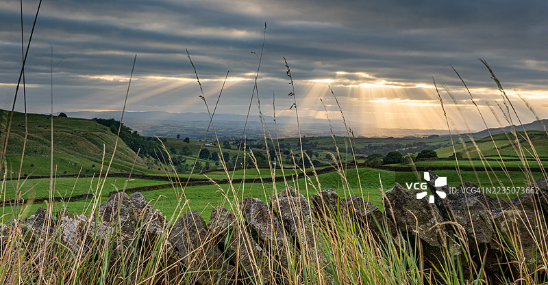 戏剧性的光线穿透云层，洒在生机勃勃的绿色农田上，前景有石墙和高草，地点：英国柯克比斯蒂芬。图片素材