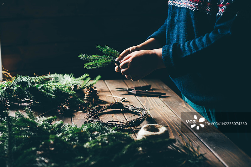 一位身穿节日北欧风格毛衣的人，正在乡村木桌上用松枝和麻绳仔细地组装圣诞花环。图片素材