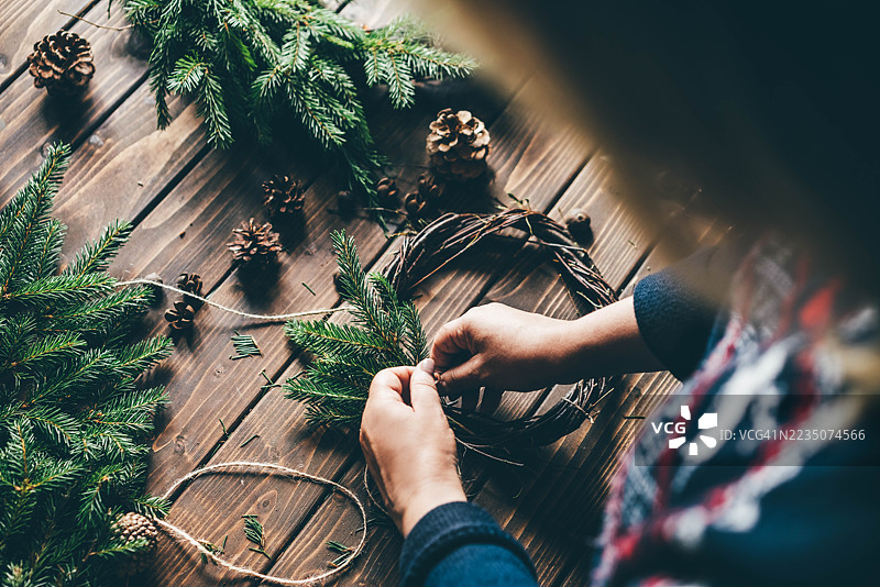一位身穿节日北欧毛衣的人，正在质朴的木桌上，用松枝和麻绳小心地组装圣诞花环。图片素材