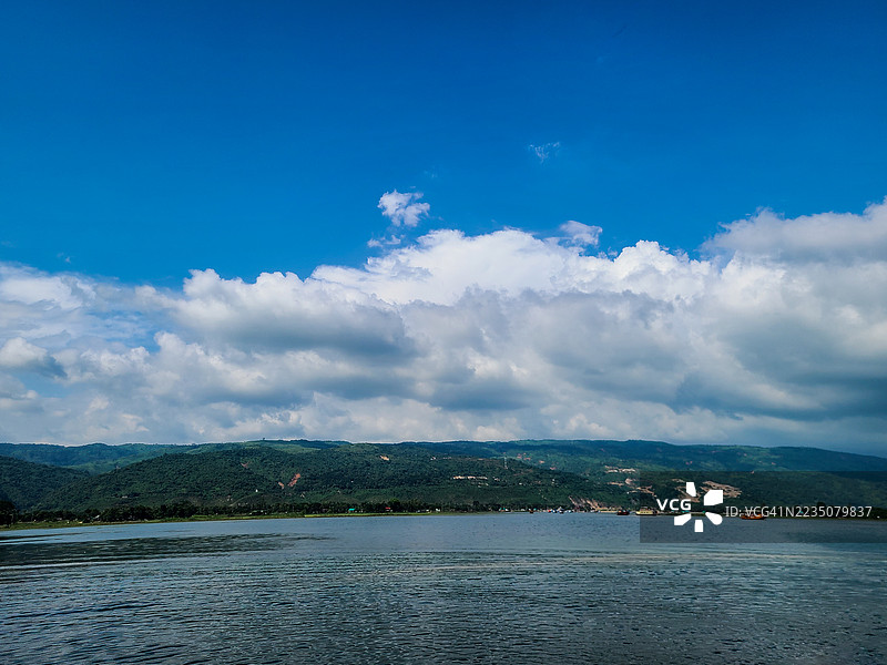 宁静湖泊的广阔景观，四周被郁郁葱葱的绿色山脉环绕，明亮的蓝天上点缀着几朵白云，孟加拉国图片素材