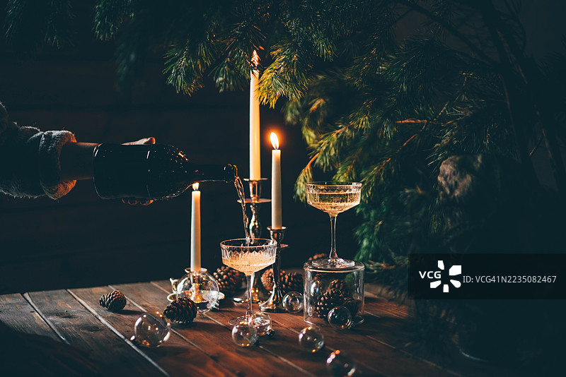 一只手将起泡酒倒入酒杯，酒杯放置在一张装饰精美的木桌上，桌上还有点燃的蜡烛、松枝和松果。图片素材