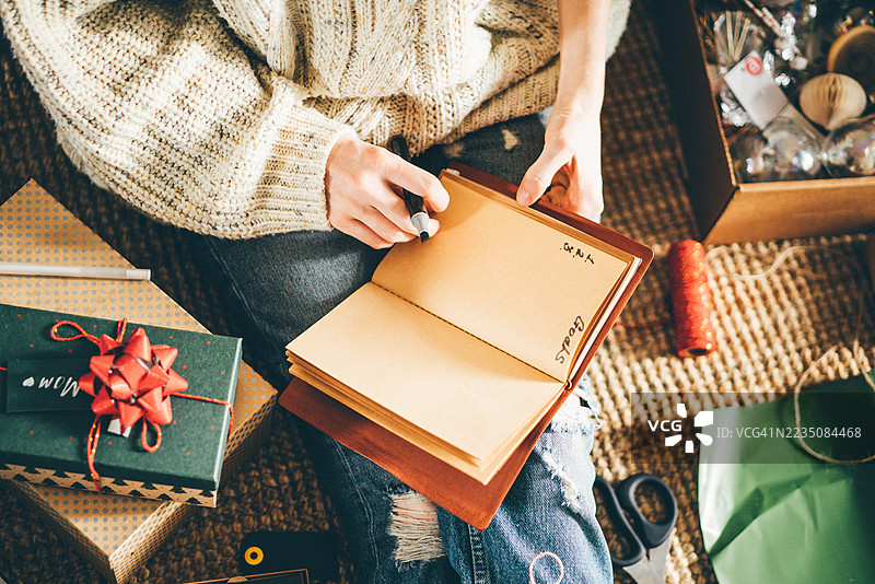 俯视角度拍摄，一名年轻女子坐在地板上，周围是圣诞包装用品和礼物，她正在笔记本上书写。图片素材