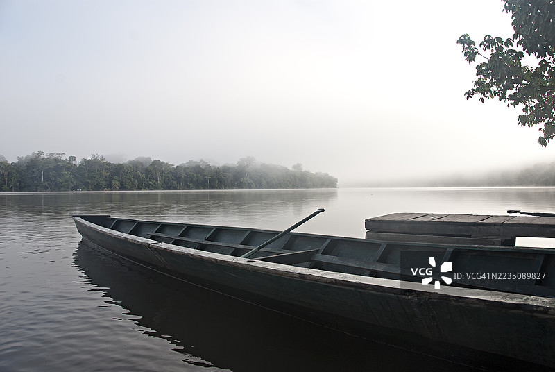 宁静的晨雾笼罩着宁静的湖泊，一只传统的木船轻轻停靠在小码头附近，周围环绕着郁郁葱葱的雾气丛林。图片素材