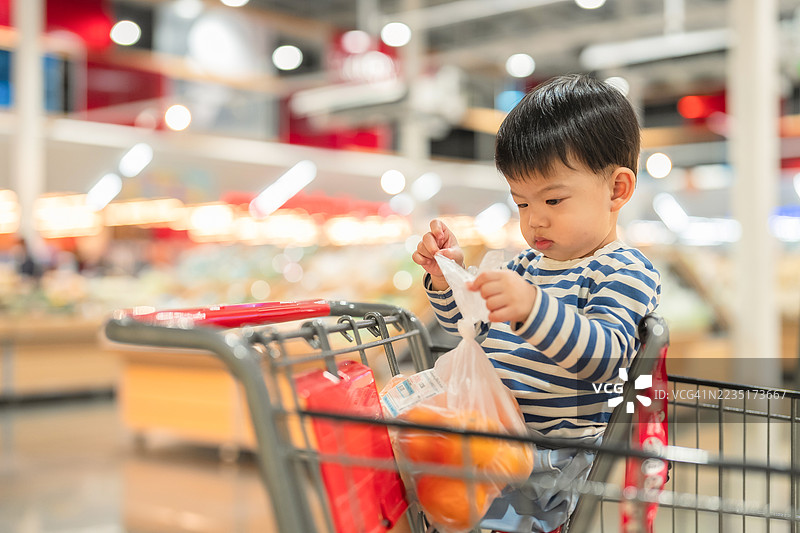 快乐的幼儿男孩坐在明亮超市的购物车里，开心地观察周围，笑得很开心。穿着休闲服的男孩散发着纯真和快乐，带来温暖和日常家庭的欢乐。图片素材