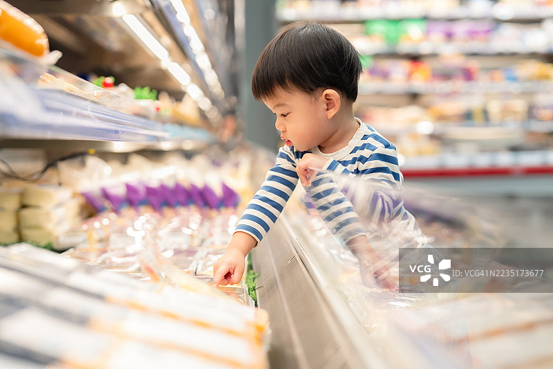 一个好奇的小男孩站在超市的冷藏区前，伸手去挑选新鲜蔬菜。这一刻捕捉了日常家庭生活中可爱的场景，展现了小男孩在购物时探索健康食品选择的情景。图片素材