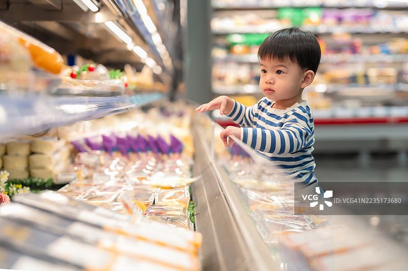 一个好奇的小男孩站在超市的冷藏区前，伸手去挑选新鲜蔬菜。这一刻捕捉了日常家庭生活中可爱的场景，展现了小男孩在购物时探索健康食品选择的情景。图片素材
