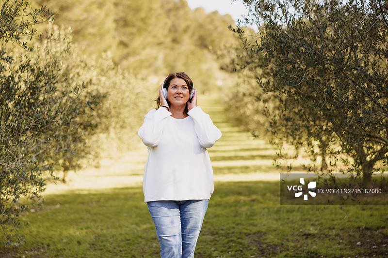 成熟的女性在橄榄树林中漫步，触摸耳机，享受音乐或播客。图片素材