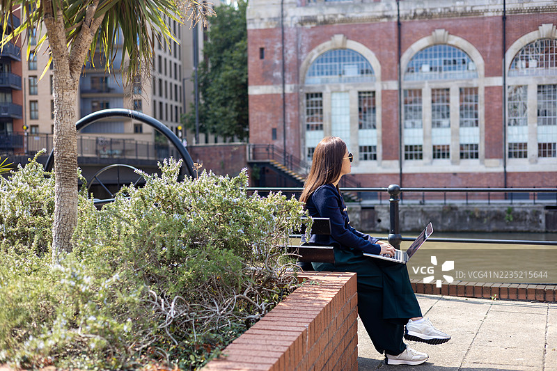 一位年轻的职业女性在英国欧洲布里斯托尔市中心的长椅上，使用笔记本电脑远程工作。图片素材