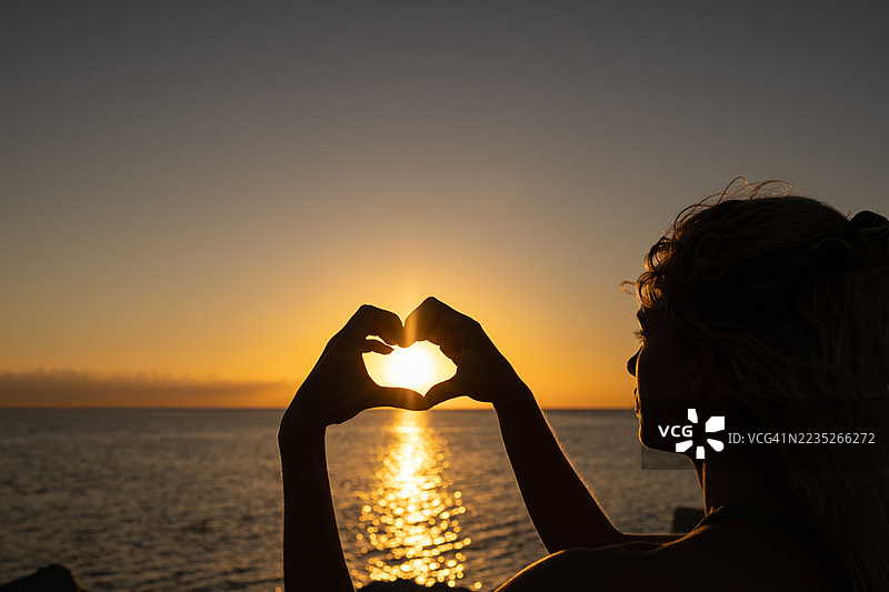 海边日落时，一位女性用双手做出心形轮廓，享受着美丽的景色图片素材