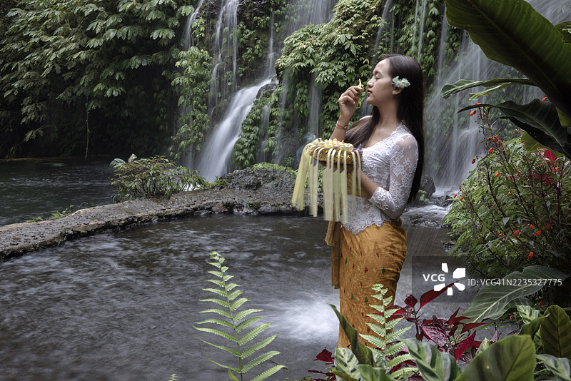 一位印尼女性身着鲜艳的传统服装，站在印尼巴厘岛的班尤瓦纳阿梅尔塔瀑布前。图片素材