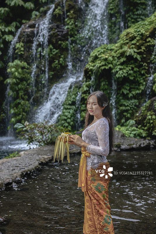 一位印尼女性身着鲜艳的传统服装，站在印尼巴厘岛的班尤瓦纳阿梅尔塔瀑布前。图片素材