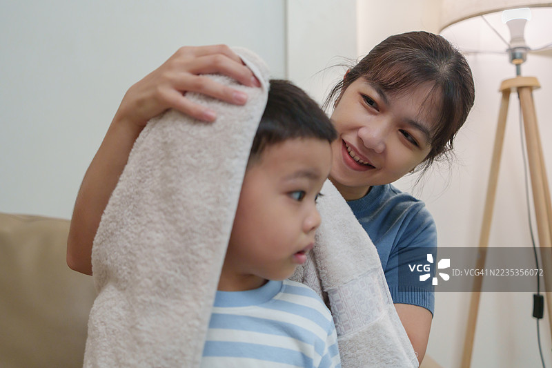 亚洲母亲在洗澡后和年幼的儿子一起坐在沙发上，用毛巾轻轻地为儿子擦干头发。图片素材
