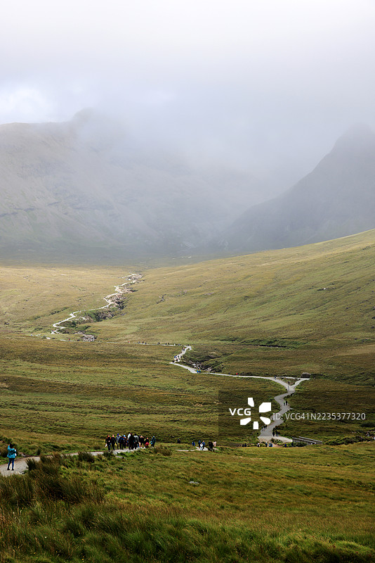 在英国苏格兰的斯凯岛，仙女池的郁郁葱葱的山谷中，一条蜿蜒的小路穿过薄雾，背景是丘陵和朦胧的山脉。图片素材