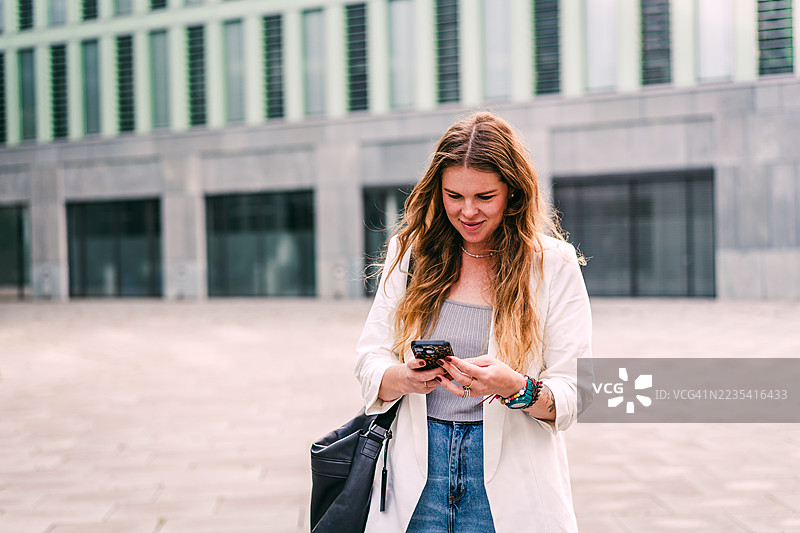自信的年轻女子在现代城市建筑外查看手机图片素材